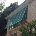 Turquoise metal mesh awning installed on a window of a textured stucco house, with green bushes and pink flowers in the foreground.